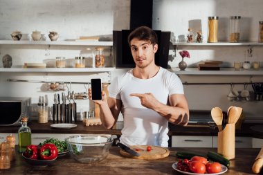mutfakta yemek yaparken boş ekran ile akıllı telefon parmak ile işaret beyaz t-shirt kaslı adam