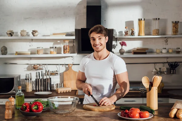 smiling muscular man in white t-shirt cutting vegetables in kitchen