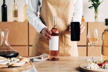 şarap şişeleri ve boş ekran ile akıllı telefon gösteren masa yakınında apron dikili Sommelier kısmi görünümü