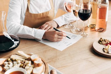 cropped view of sommelier in apron sitting at table and writing in wine tasting document clipart