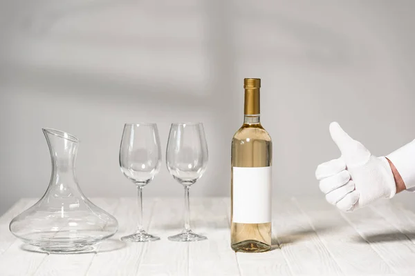 partial view of waiter in white glove showing thumb up near table with bottle of wine, wine glasses and jug