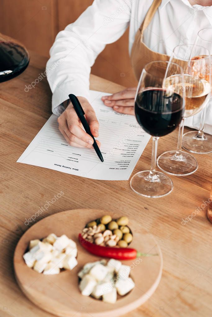 cropped view of sommelier in apron sitting at table and writing in wine tasting document