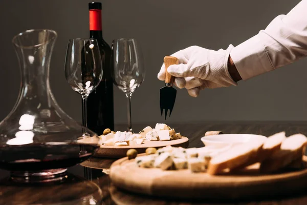 cropped view of waiter in white glove holding cheese fork near table with food and wine