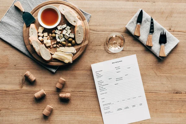 top view of wine tasting document, wine glass, food, cutlery and corks on wooden surface