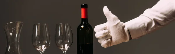 panoramic shot of waiter in white glove showing thumb up near bottle of wine and wine glasses