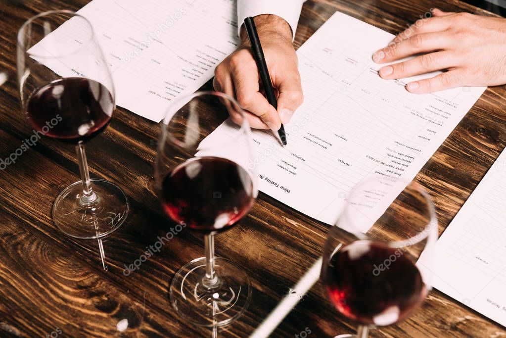 cropped view of sommelier writing in documents at table with wine glasses