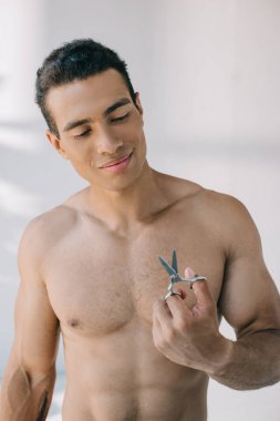 muscular mixed race man holding scissors and smiling while looking on it clipart