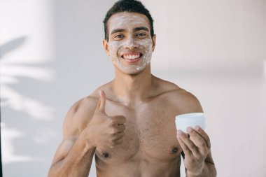 handsome mixed race man with cosmetic cream on face holding container and showing thumb up while smiling and looing at camera clipart