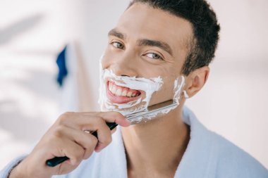 handsome young man in bathrobe shaving face with razor blade, smiling and looking at camera clipart