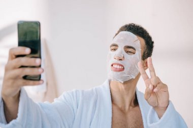 young man in bathrobe with cosmetic mask on face taking selfie on smartphone and showing victory sign
