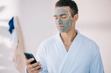 yüz ve bornoz tutan akıllı telefonda kil maskesi ile yakışıklı adam 
