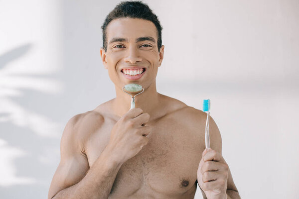 красивый смешанной расы мужчина массирует лицо нефритовым роликом и держит зубную щетку, глядя в камеру
