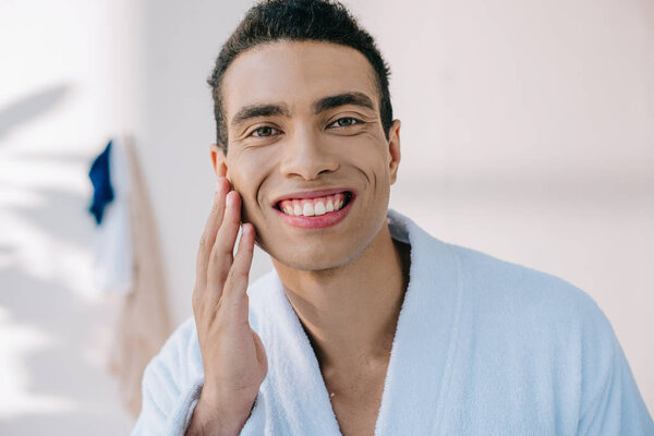handsome man in bathrobe touching face while smiling and looking at camera