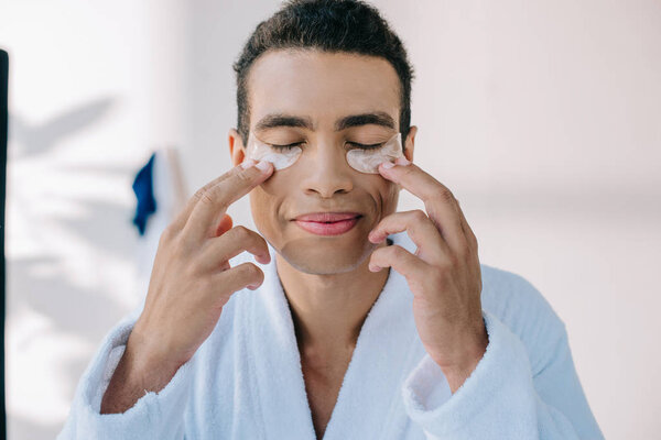 young man putting patches for eyes and smiling while standing in bathrobe with closed eyes