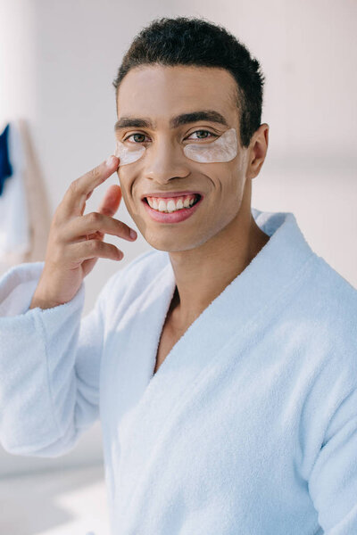 portrait shot of handsome man in bathrobe touching patches, smiling and looking at camera