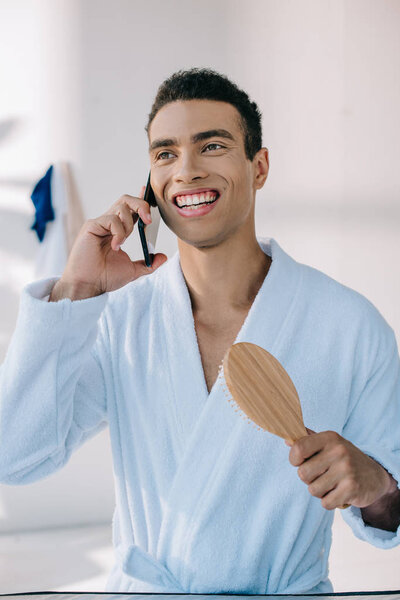 handsome man in bathrobe holding hairbrush and talking on smartphone while smiling and looking away