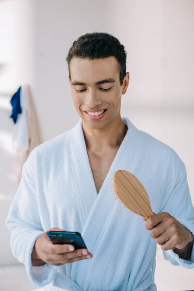 handsome man in bathrobe holding hairbrush and smartphone while smiling and looking away