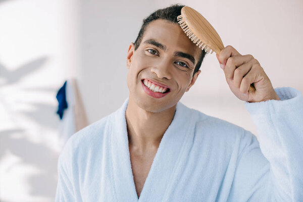 handsome young man in bathrobe combing hair with hairbrush while amiling and looking at camera