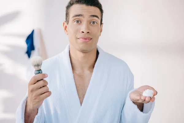 Joven Disgustado Albornoz Sosteniendo Cepillo Afeitar Crema Afeitar Mirando Cámara — Foto de Stock