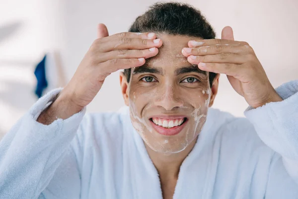 handsome man in bathrobe washing face with facial foam and smiling while looking at camera