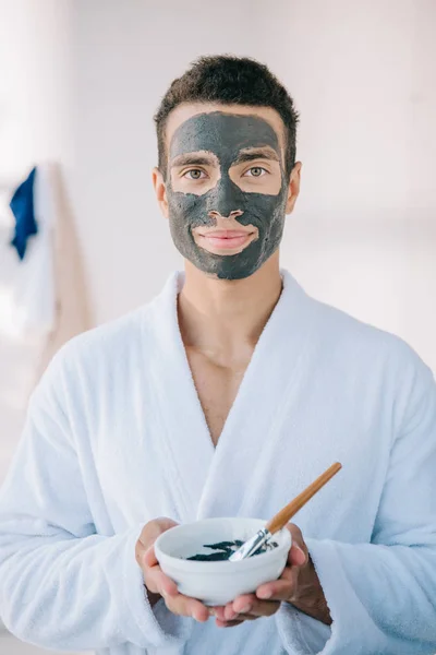 Joven Guapo Albornoz Sosteniendo Tazón Con Mascarilla Cepillo Mientras Mira —  Fotos de Stock