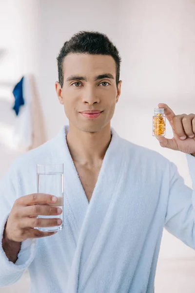 英俊的男人在浴袍拿着瓶子与维生素和玻璃与水 看着相机 — 图库照片