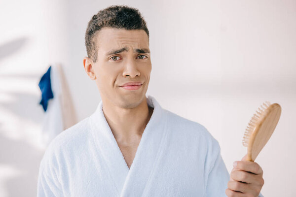 disappointed young man in bathrobe holding hairbrush, looking at camera and making grimace