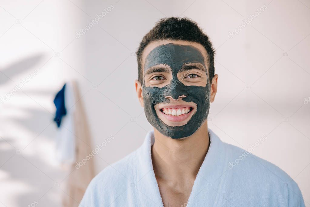 handsome young man in bathrobe and with grey clay mask on face smiling and looking at camera