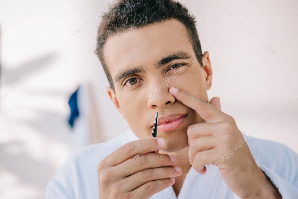 handsome young man plucking nose with tweezer and looking at camera