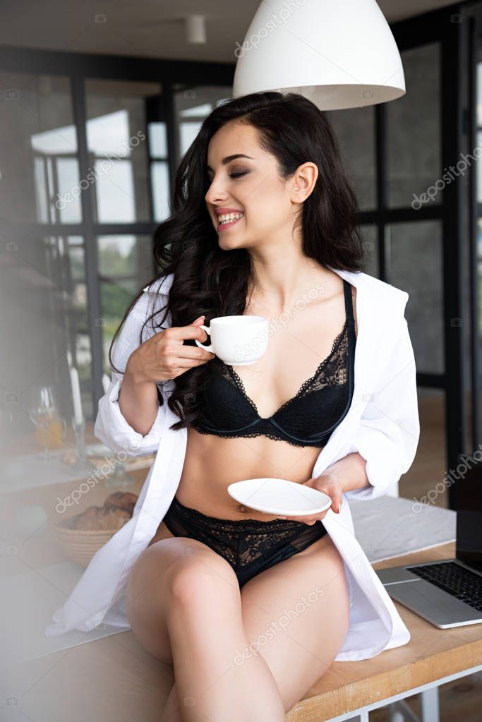 sexy girl in black underwear and white shirt drinking coffee with closed eyes while sitting on table in kitchen 