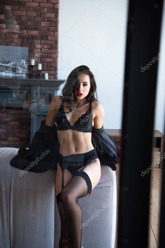 front view of sexy girl in black underwear with jacket sitting on sofa in living room and looking at camera