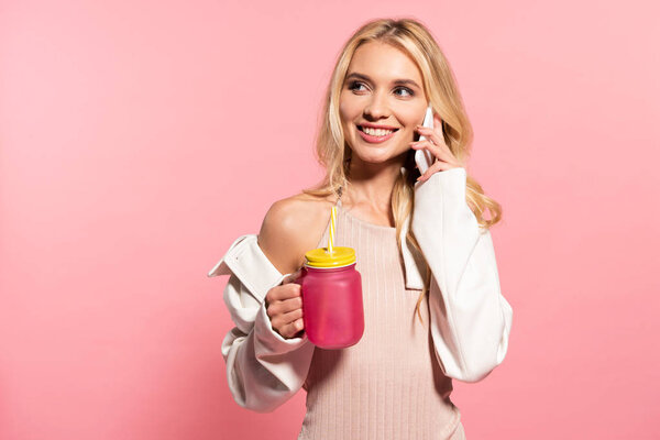 beautiful blonde girl holding beverage and talking on smartphone on pink