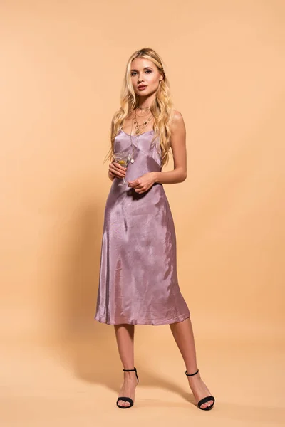 优雅的金发女郎在紫色的丝色连衣裙和项链举行鸡尾酒米色 — 图库照片