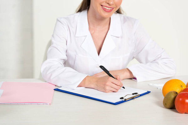 cropped view of smiling dietitian in white coat at workplace writing in clipboard with fruits on table