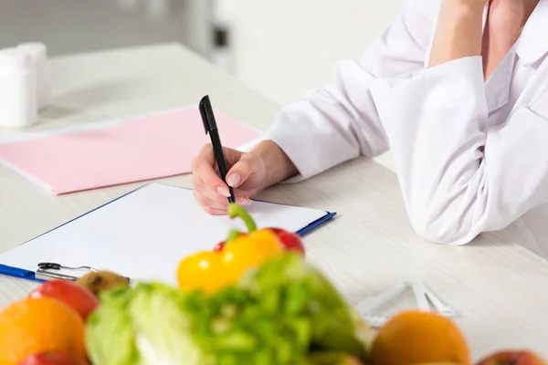 テーブルの上に果物や野菜と職場のクリップボードに書き込み白いコートの栄養士の部分的なビュー — ストック写真