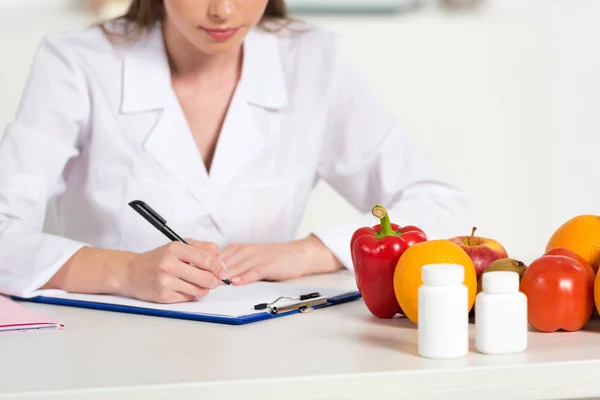 テーブルの上に錠剤 果物や野菜と職場でクリップボードに書き込み白いコートでトリミングビュー栄養士 — ストック写真