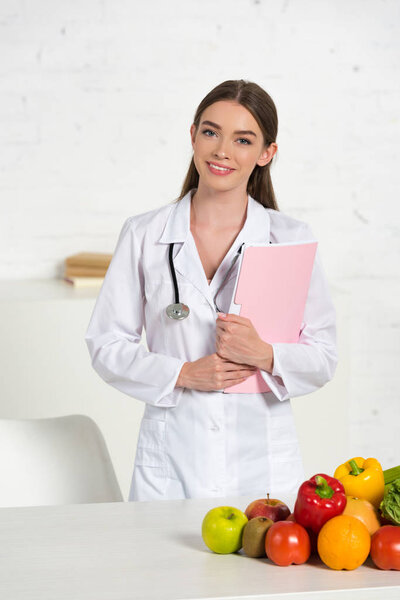 смайлик привлекательный диетолог в белом пальто с оборудованием холдинга папку рядом со столом со свежими фруктами и овощами

