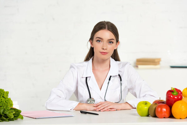 front view of dietitian in white coat with equipment sitting at workplace with fruits and vegetables on table