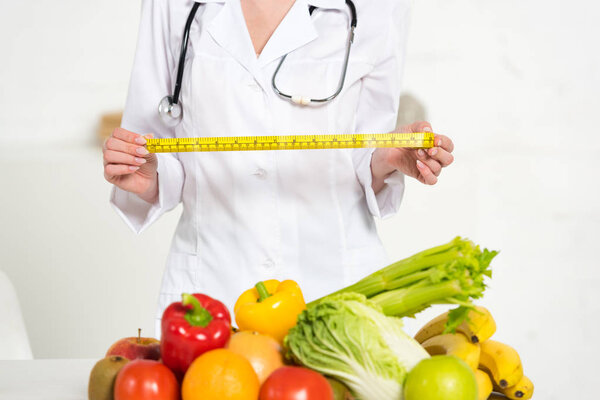 обрезанный вид диетолога в белом пальто проведение меры ленты рядом со свежими фруктами и овощами
