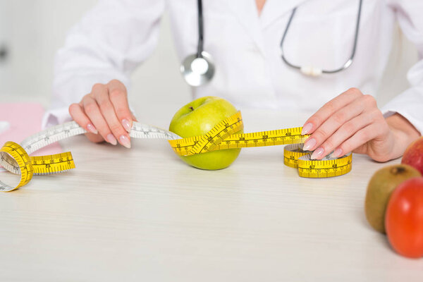 cropped view of dietitian in white coat holding measure tape at table with fruits