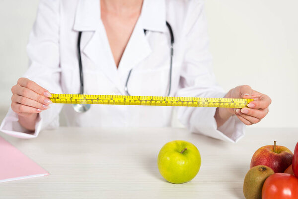 cropped view of dietitian in white coat holding measure tape and fresh food on table
