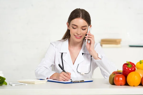Smiling Dietitian White Coat Talking Smartphone Writing Clipboard Workplace — Stock Photo, Image