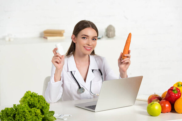 smiling dietitian in white coat holding pills and carrot near laptop