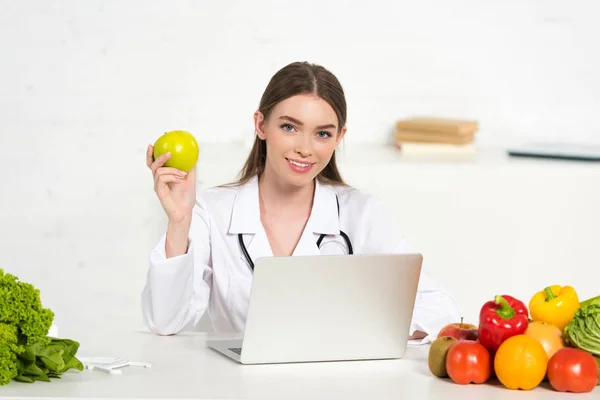 Uśmiechnięty Dietetyk Białym Płaszczu Trzymając Jabłko Miejscu Pracy Laptopem — Zdjęcie stockowe