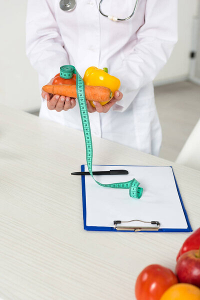 cropped view of dietitian in white coat holding vegetables and measure tape at workplace