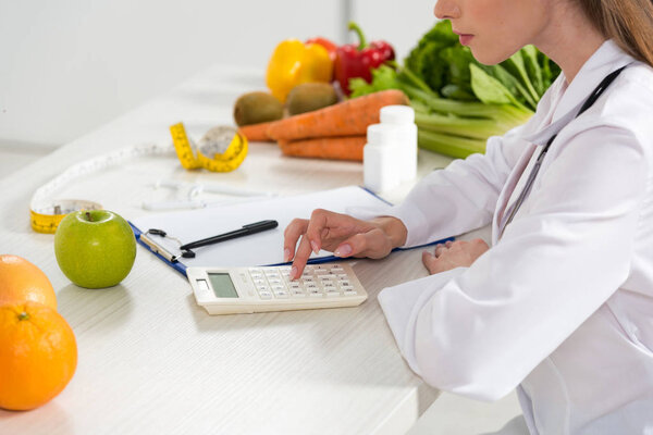 обрезанный вид диетолога в белом халате с помощью калькулятора на рабочем месте
