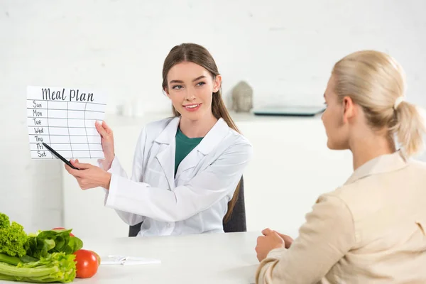 微笑营养师在白色外套举行膳食计划和病人在餐桌上 — 图库照片