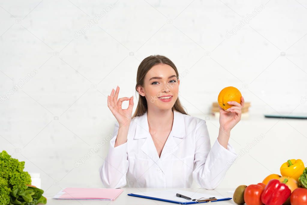 smiling dietitian in white coat holding orange and showing okay sign at workplace