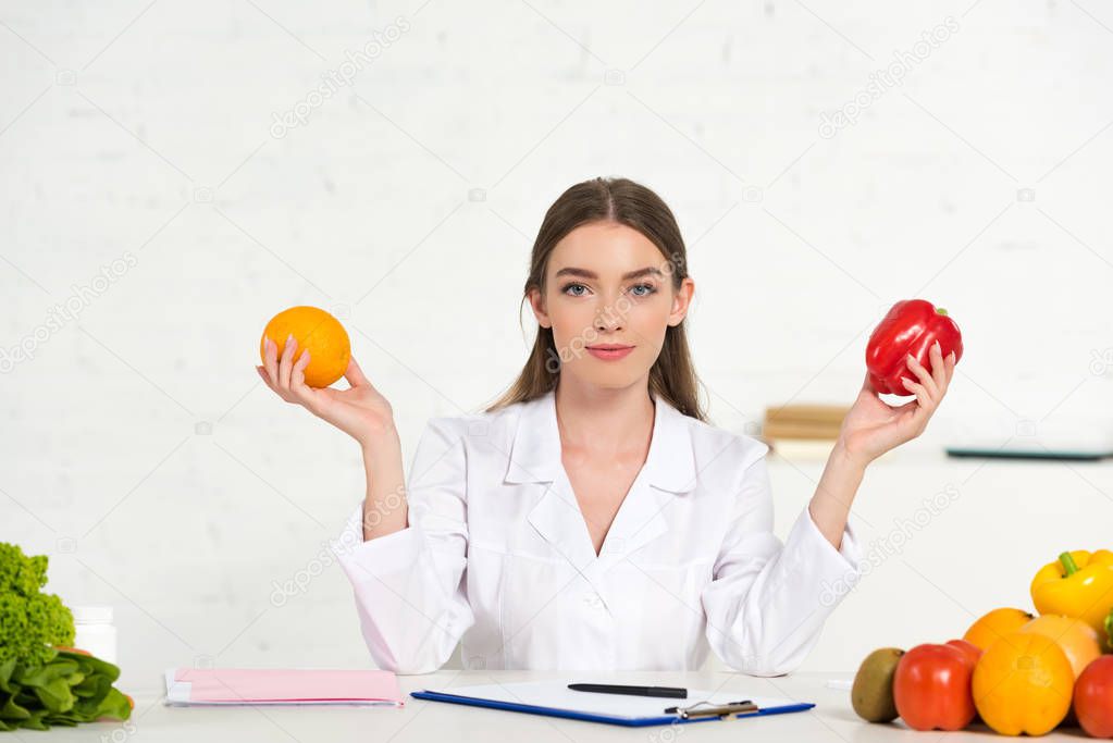 front view of dietitian in white coat holding orange and bell pepper at workplace