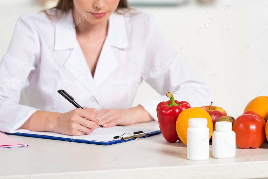 cropped view dietitian in white coat writing in clipboard at workplace with pills, fruits and vegetables on table